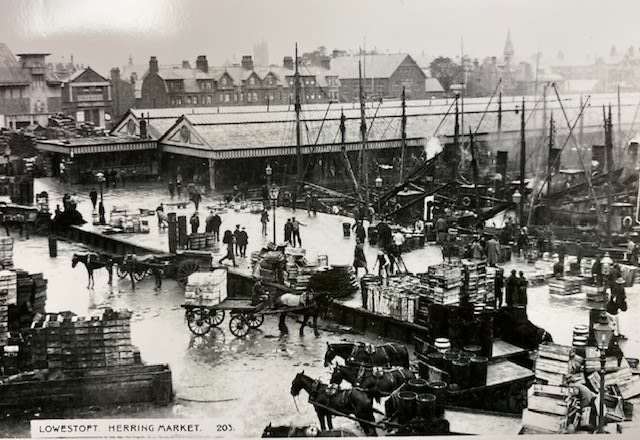 A greyscale image showing the harbour village of Lowestoft. The harbour is a hive of activity with ships moored in the background of the image (some powered by steam, others by wind). Moving inwards, approximately 100 people can be seen completing various activities including loading and unloading cargo onto a horse and trailer; stacking fishing crates onto various piles in the harbour; offloading the vessels using their hands; and others are either chatting or walking briskly to their next task. A large warehouse facility appears on the right back corner of the photograph and it is made up of pillars with two triangular roofs and a forward part on the second half of the warehouse that has a flat roof. Gutters on the roof are viewable. 